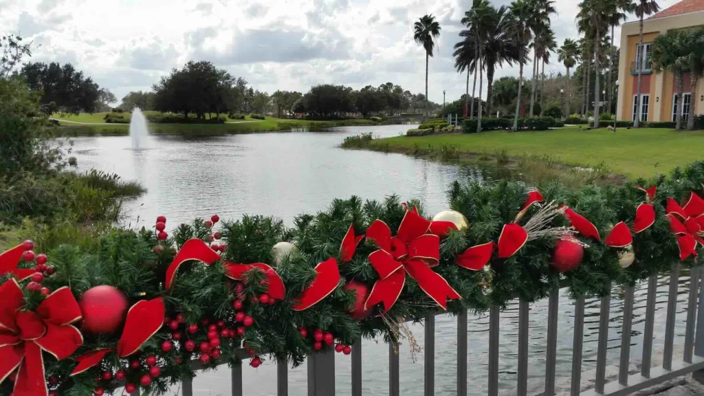 Festive holiday garland with red bows and ornaments decorates bridge railing overlooking pond with palm trees - Lindholm Exteriors