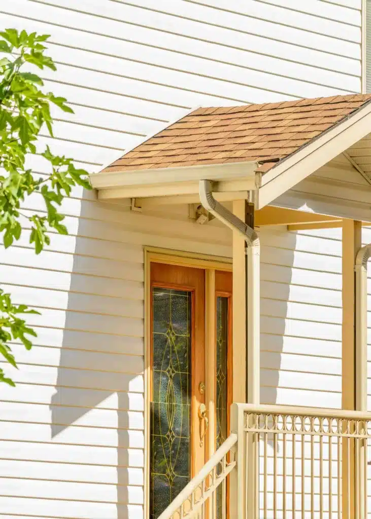 White siding and beige railing, next to wooden front door with decorative glass under small shingled porch, leafy branch - Lindholm Exteriors